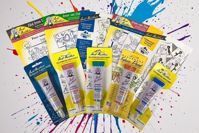 5 tubes of Aunt Martha’s brand fabric paint in various colors, arranged with their packaging on a painted splash background.
