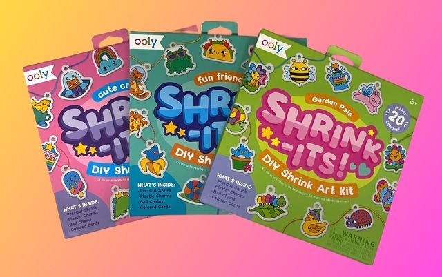 Three OOLY Shrink-Its! DIY shrink art kits in pink, blue, and green featuring cartoon designs against a colorful background.