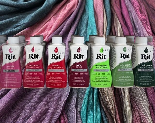 A row of seven Rit liquid dye bottles in shades of red, pink, and green, arranged in front of matching colored fabric.