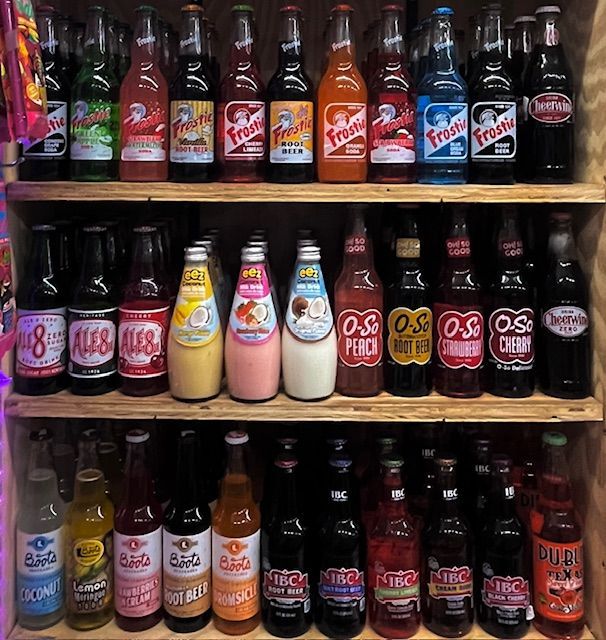 Three shelves filled with various brands and flavors of glass-bottled craft sodas arranged in rows.