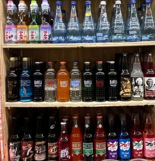 Three wooden shelves stocked with various glass-bottled sodas in a wide range of colors and brand labels.
