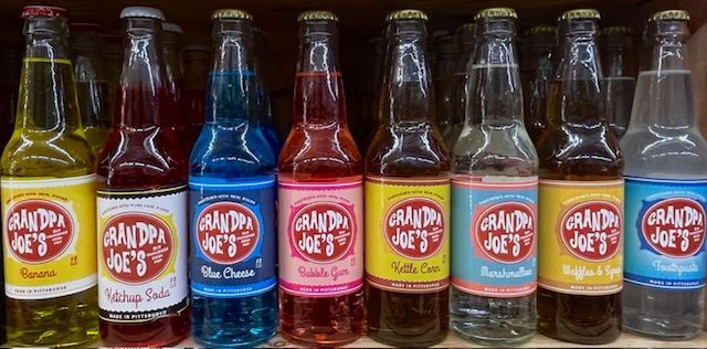 A shelf displaying eight colorful bottles of Grandpa Joe’s novelty sodas, featuring flavors like Ketchup and Blue Cheese.