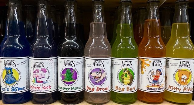 Seven glass bottles of novelty sodas with colorful liquids and labels with names like Toxic Slime and Bug Barf on a shelf.