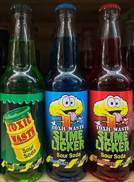 Three bottles of Toxic Waste Slime Licker sour soda in green apple, blue razz, and strawberry flavors on a store shelf.