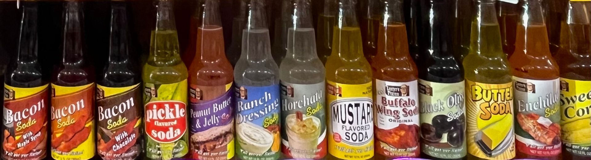 A row of unusual novelty soda flavors in glass bottles, including bacon, pickle, ranch, mustard, and corn-themed options.