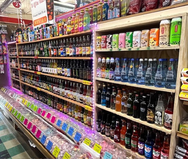A store shelf filled with various bottled sodas, canned drinks, and colorful candy in bins underneath.