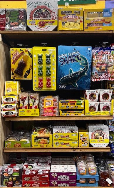 Retail store shelves stocked with various novelty candy boxes, including gummy sharks, pizza, and soda-shaped treats.