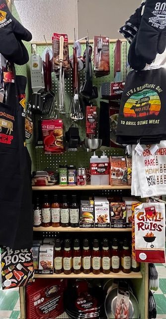 A retail display shelf filled with grilling supplies, including tools, spice rubs, sauces, and themed kitchen linens.