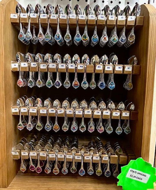 A wooden rack displays rows of souvenir spoons with colorful emblem-decorated handles, with a green price tag on the side.
