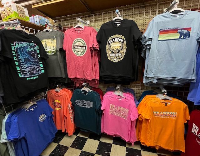 A rack of colorful T-shirts featuring various Brandon, Missouri designs, displayed in a retail shop with checkered floors.