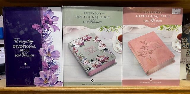 Three Everyday Devotional Bibles for Women in purple, floral, and pink designs, displayed on a wooden shelf.