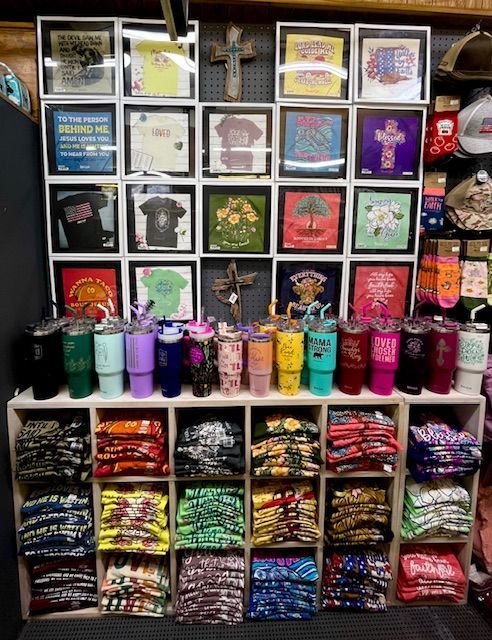 A retail display featuring framed t-shirt designs on a wall above a shelf of colorful tumblers and folded t-shirts.