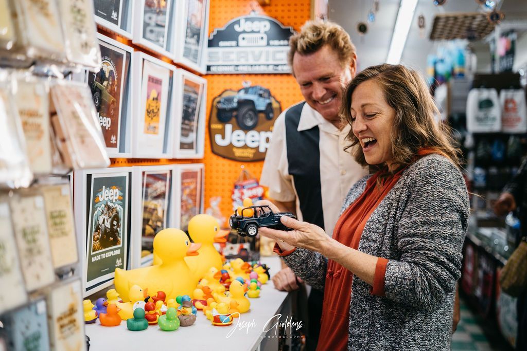 A woman smiles while holding a toy jeep, standing with a man in a shop filled with Jeep-themed merchandise and rubber ducks.