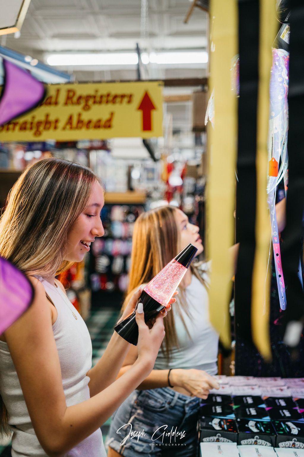 Two people examine a pink lava lamp in a retail store with a sign pointing toward registers.