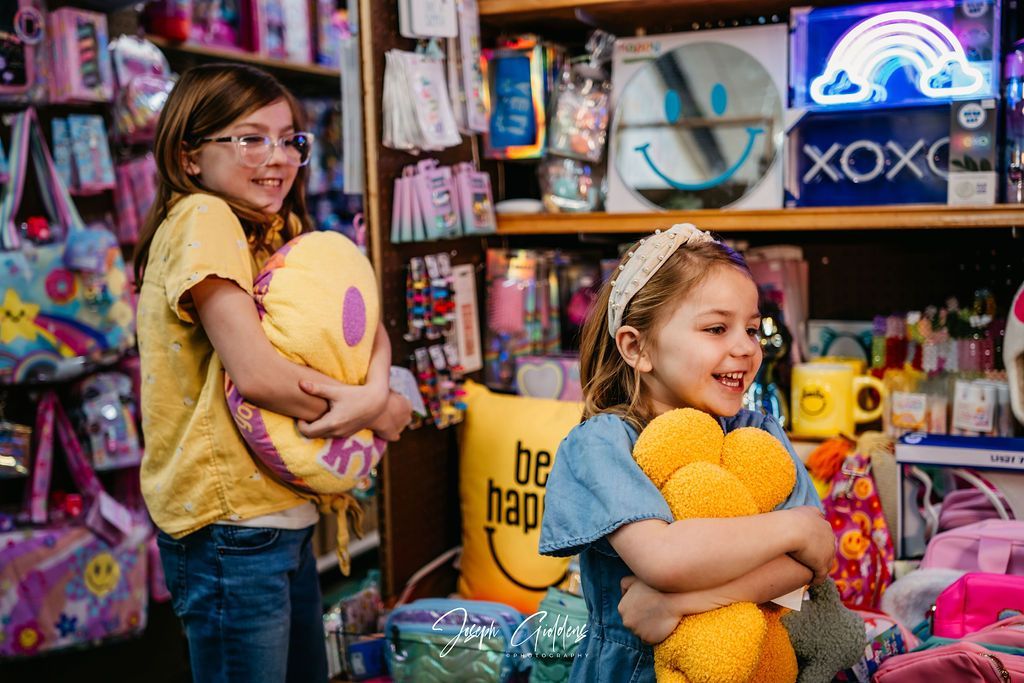 Two children in a store hug yellow plush toys, smiling amidst shelves filled with colorful accessories and gifts.