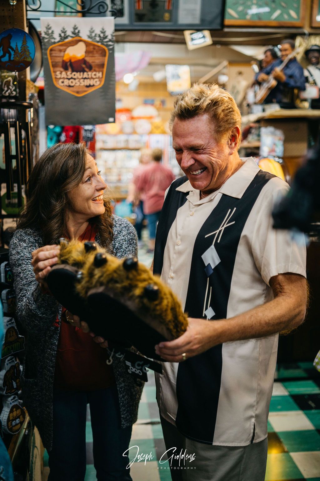 Two people smiling while inspecting a fuzzy, brown novelty item in a gift shop under a Bigfoot Crossing sign.