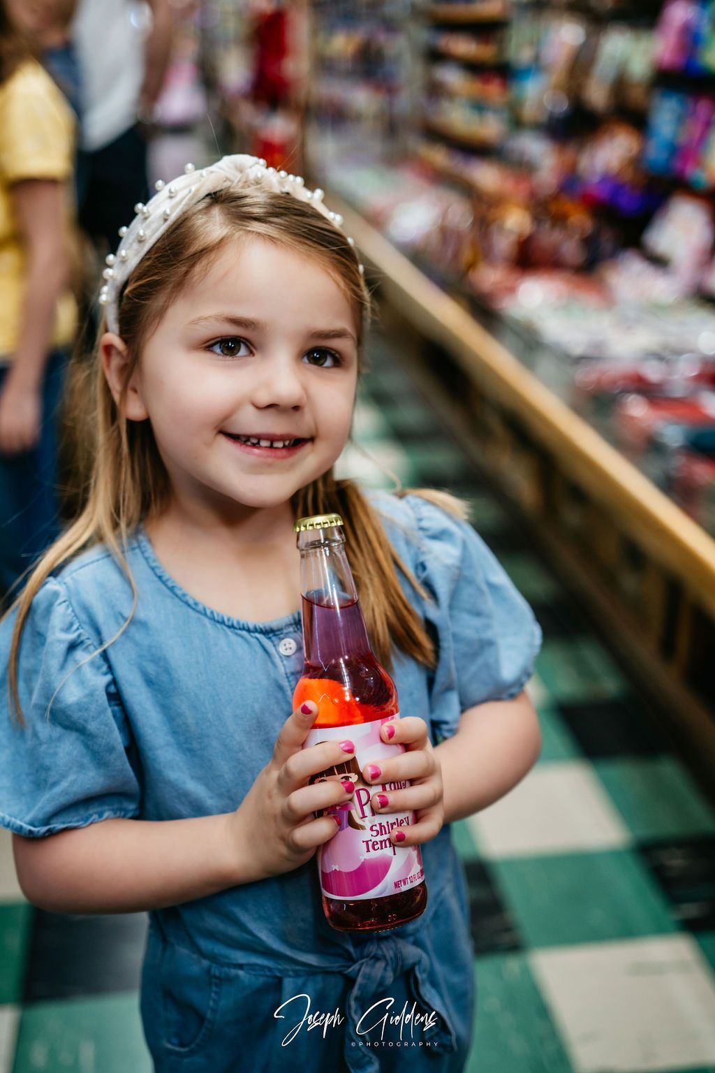 A smiling child with a beaded headband holds a bottle of pink soda in a store with a checkered floor.