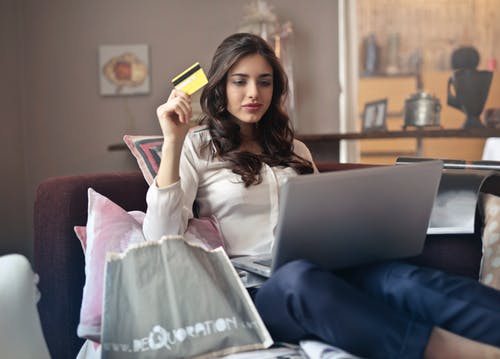 Uma mulher está sentada em um sofá segurando um cartão de crédito e usando um laptop.