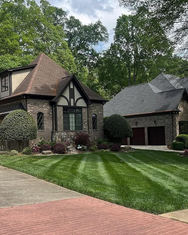 Brick house exterior with green shutters, landscaped shrubs, and a concrete walkway beside a lawn