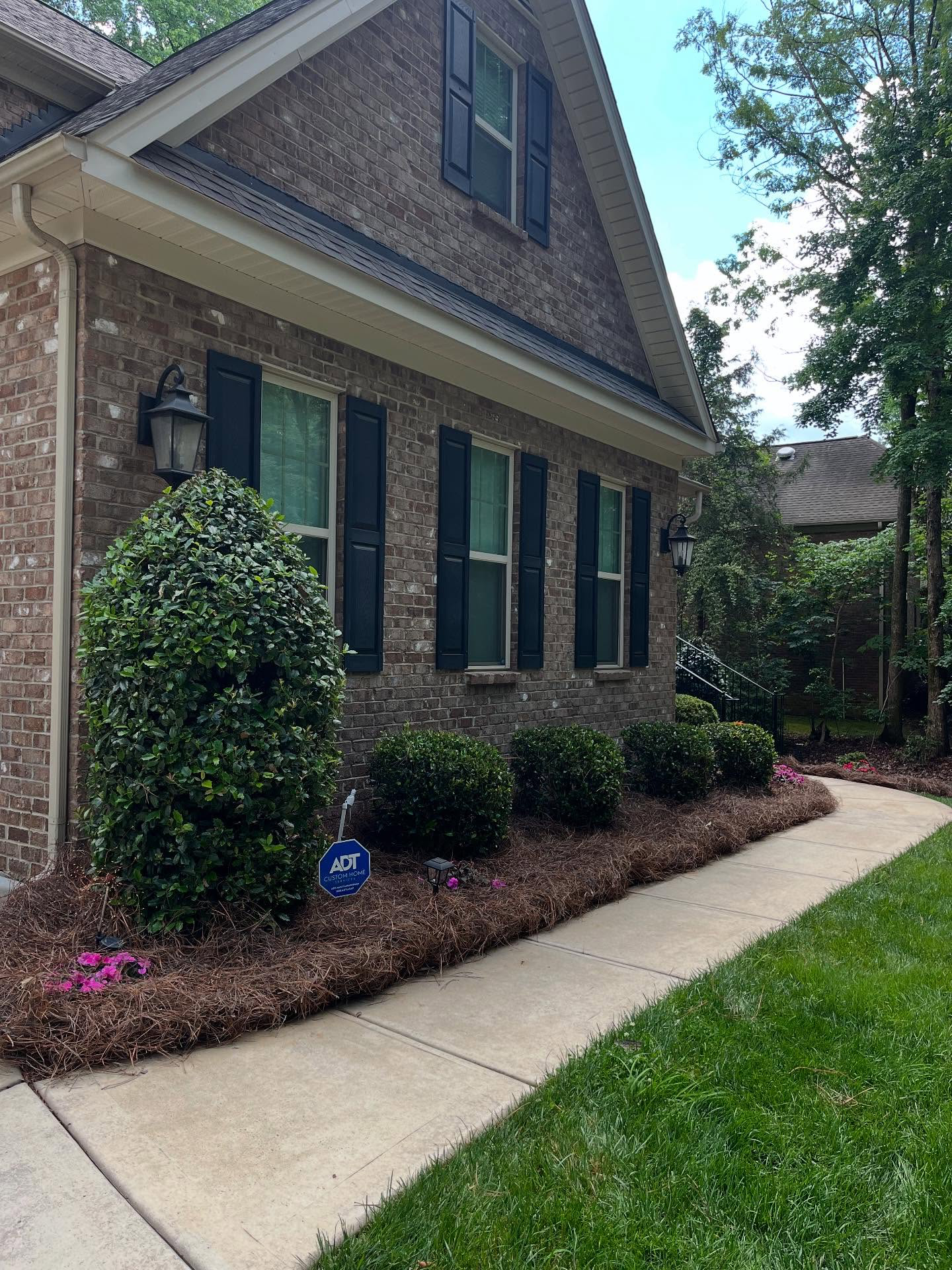 Brick house exterior with green shutters, landscaped shrubs, and a concrete walkway beside a lawn