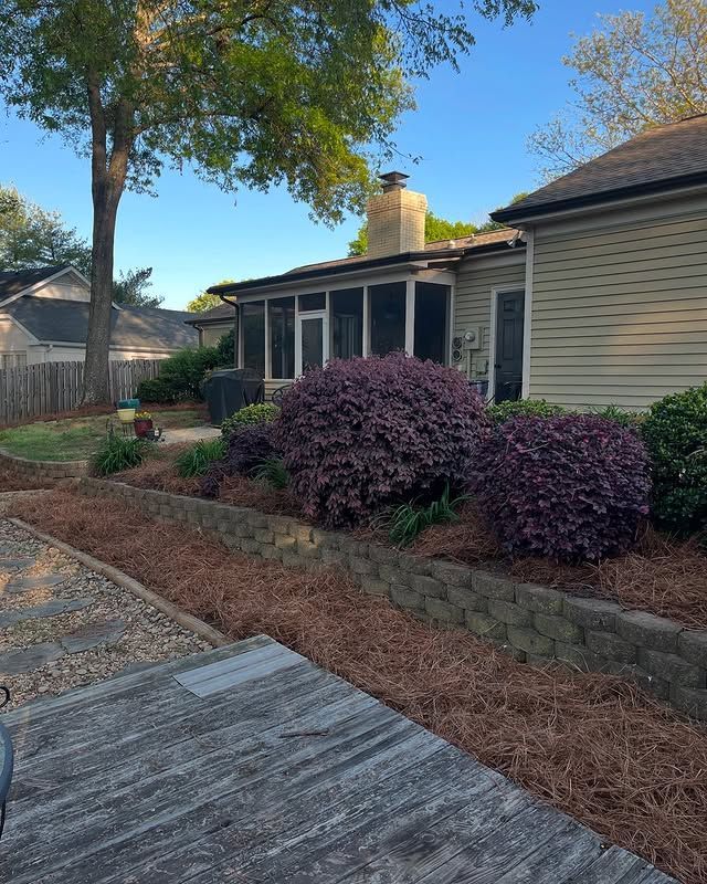 Brick house exterior with green shutters, landscaped shrubs, and a concrete walkway beside a lawn
