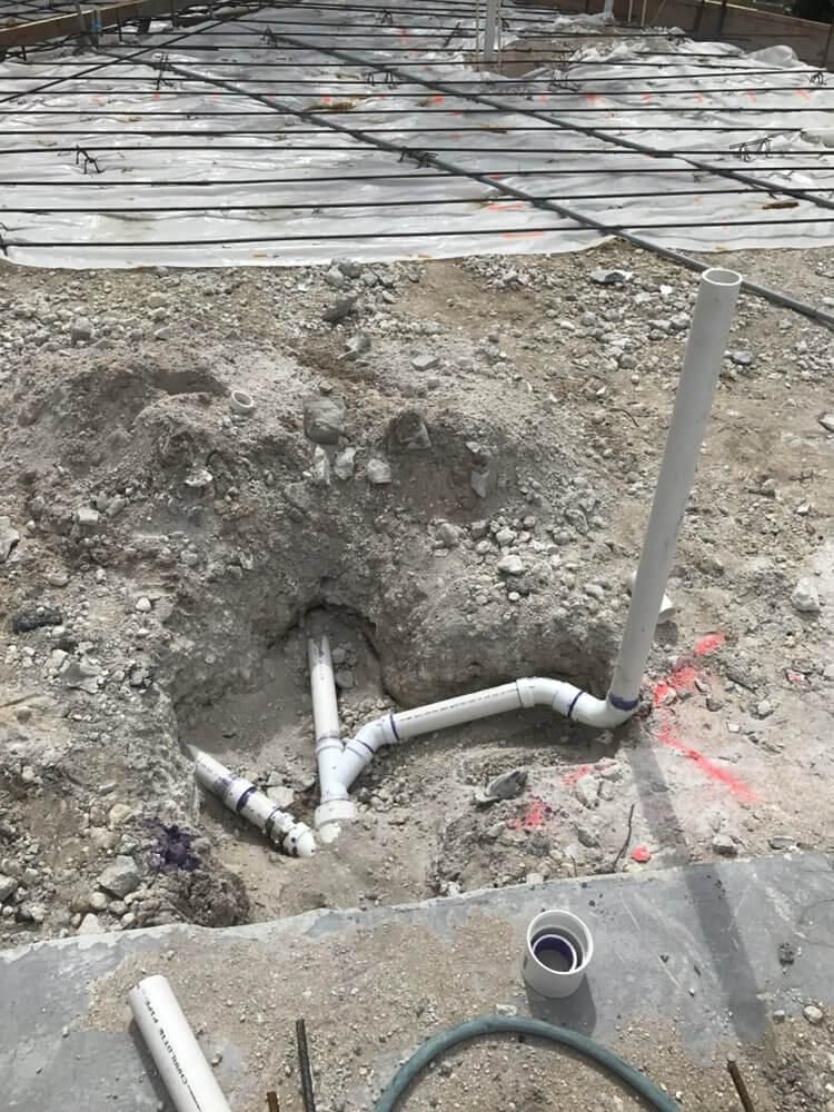 Broken white pipes in a dirt excavation beside a concrete walkway and rebar grid.