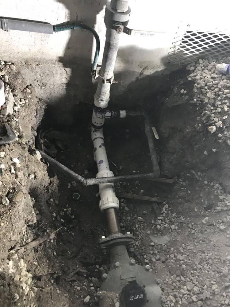 Open trench exposing underground pipes and fittings beside a wall, with dirt and gravel around them