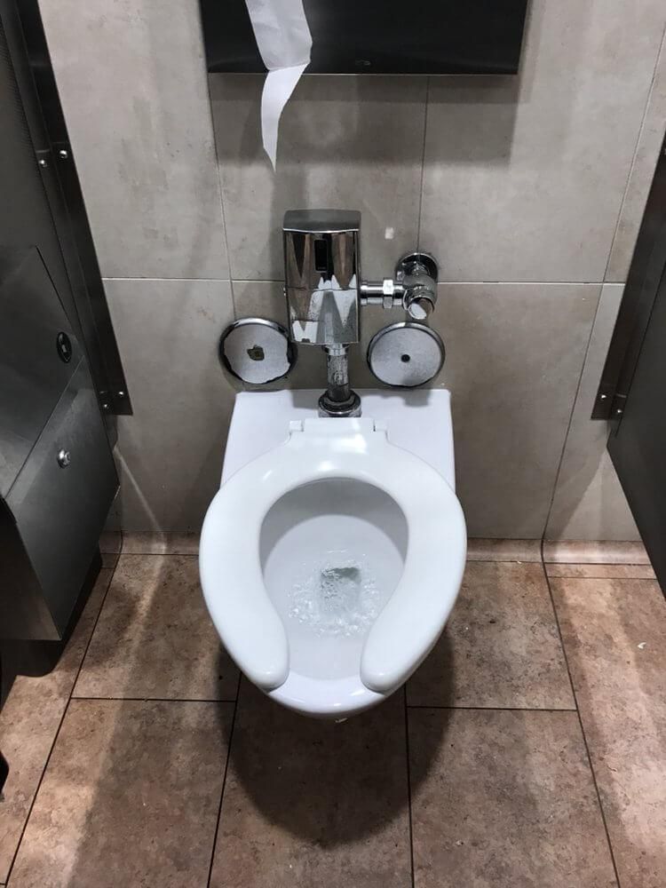 Public restroom toilet with metal bidet sprayer and dual toilet paper holders against tiled wall