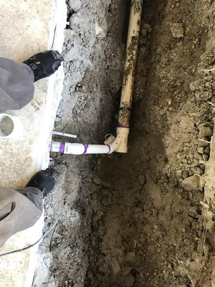 Hands standing by an open trench with exposed plumbing pipes and muddy soil.