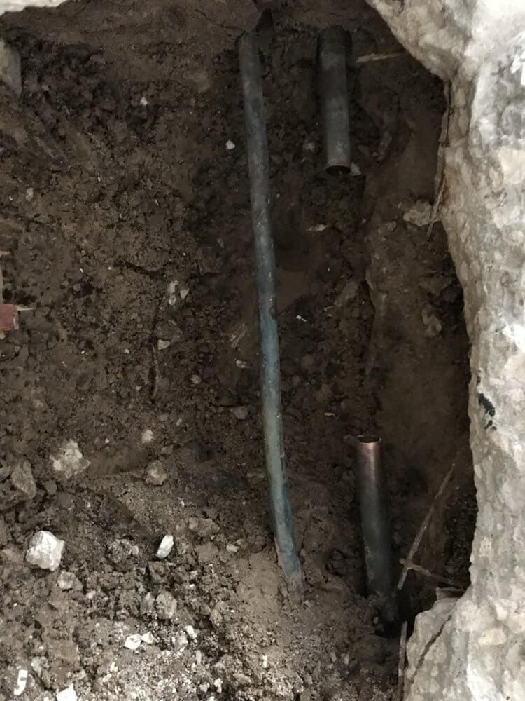 Narrow dirt trench with exposed pipes and cables beside a stone wall
