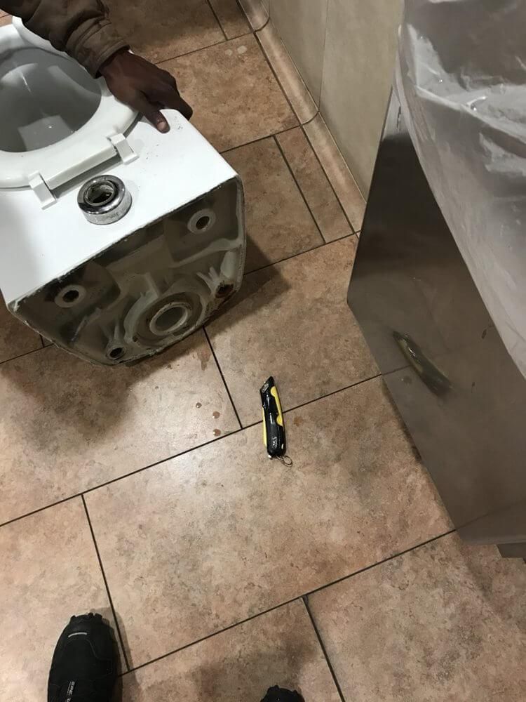 Person working on a white appliance with a tool on a tiled floor, next to another large appliance.