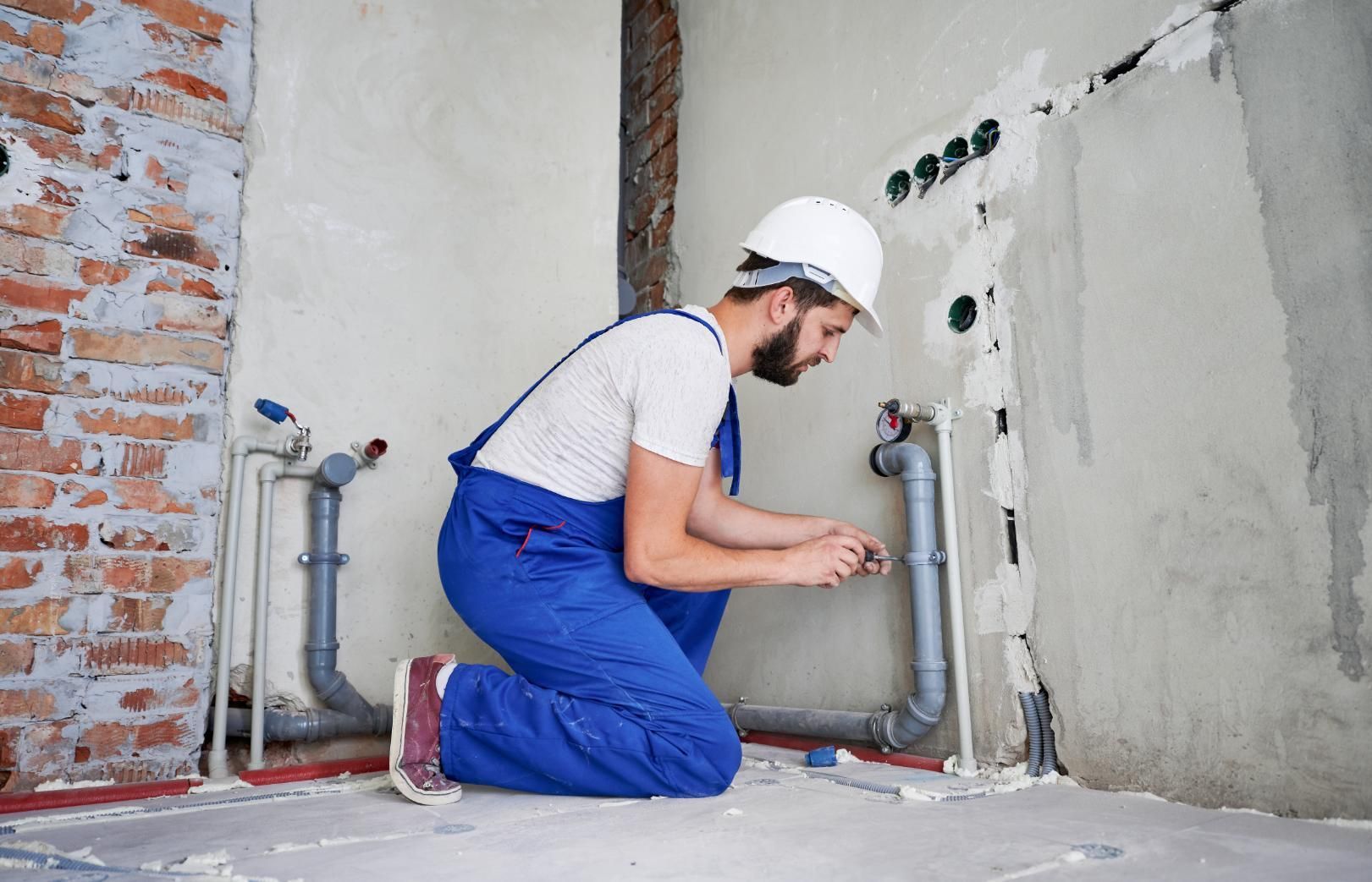 Worker in blue overalls kneeling to repair pipes in a partially finished wall