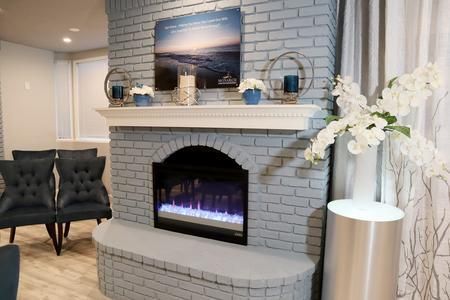A grey brick fireplace with an electric insert, white mantel, beach artwork, blue decor, and side chairs in a bright room.