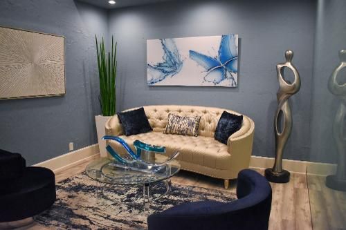 A modern lobby features a tufted beige sofa, blue chairs, a glass coffee table, abstract art, and a tall metal sculpture.