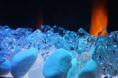 A close-up of decorative, translucent blue crystals piled on top of smooth white stones, illuminated by orange fire light.
