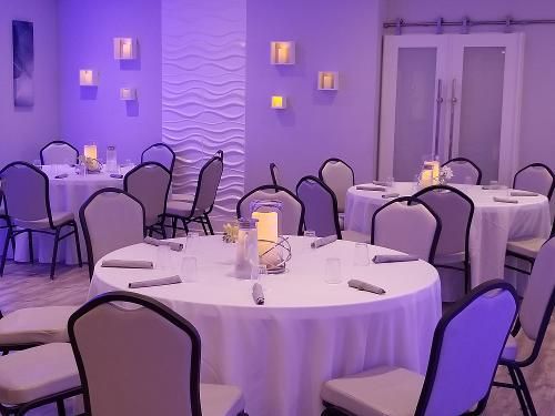 A room set for a formal event with white-clothed round tables, gray chairs, and soft purple ambient wall lighting.
