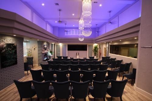 An empty event hall featuring rows of dark chairs facing a central stage, accented by purple cove lighting and chandeliers.