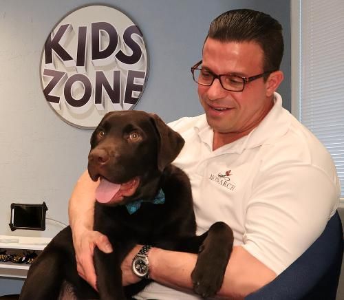 A person wearing glasses and a white polo shirt holds a dark-coated Labrador puppy wearing a blue bowtie in a Kids Zone.