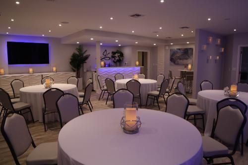 A softly lit event space with round tables covered in white cloths, grey chairs, a central bar, and a television screen.