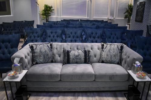 A grey tufted couch sits in front of rows of blue tufted chairs in a well-lit, contemporary room with side tables.