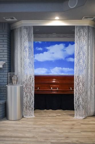 A polished wooden casket sits on a stand against a wall with a blue sky and clouds mural, framed by sheer white curtains.