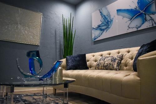 A tufted beige sofa sits in a blue-walled room with abstract art, a glass coffee table, and a tall potted plant.