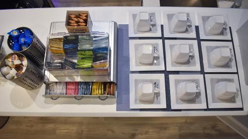 A coffee station on a white counter featuring a tea selection, condiments, and a grid of nine white ceramic mugs.