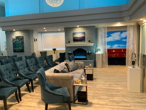 A funeral home parlor featuring rows of teal armchairs, a central sofa, a stone fireplace, and a backdrop of blue sky.