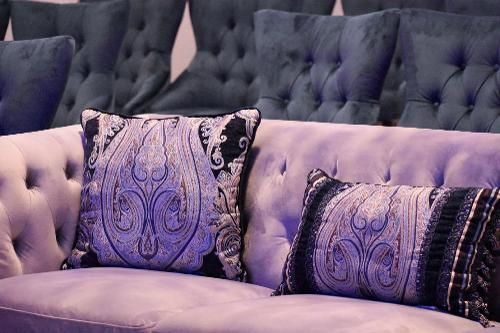 A light purple tufted sofa features two decorative throw pillows with black and purple intricate patterns.