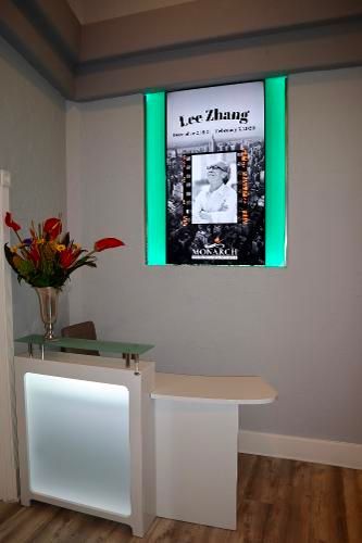 A modern reception desk with a glowing base and flower vase sits below a glowing digital memorial display for Lee Zhang.