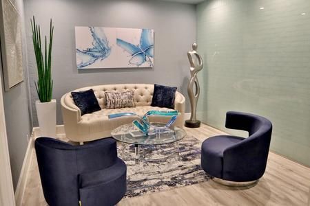 A stylish waiting area features a tufted beige sofa, two navy blue chairs, a glass coffee table, and abstract wall art.