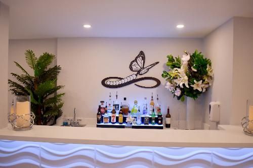 A lit, modern home bar with a butterfly wall art piece, shelves of assorted bottles, and greenery on both sides.