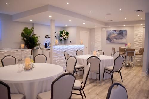 Modern, well-lit event space with round tables, white tablecloths, chairs, and a bar area with white textured fronts.