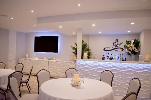 An elegant room featuring round tables with white tablecloths, chairs, and a modern bar with wavy, backlit panels.